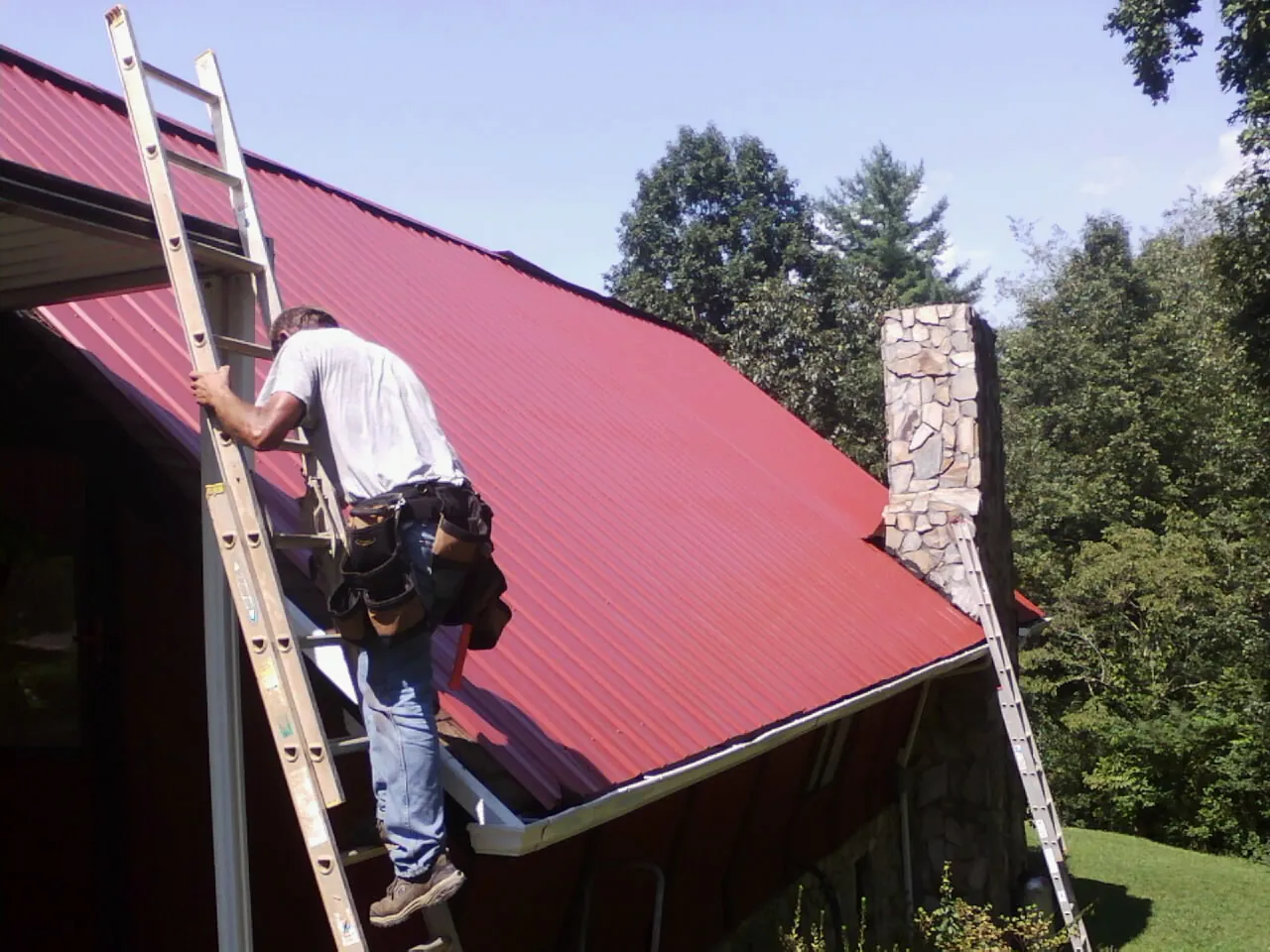 Roofer on a ladder beside a red metal roof and stone chimney — Soffit & Fascia Repair by Roofing Brothers in South Cle Elum